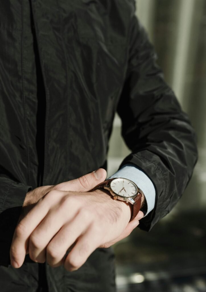 A man in a black coat adjusts his leather wristwatch outdoors, focusing on time management.