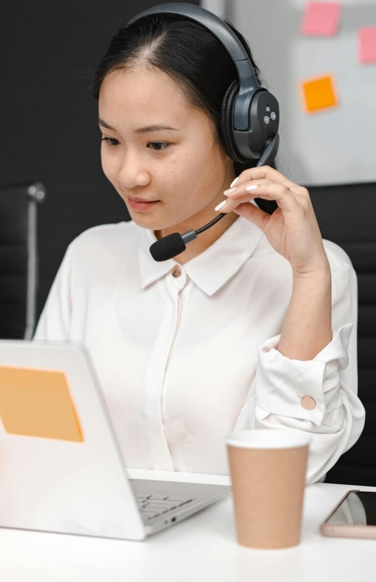 Asian woman using a laptop and headset for customer support in an office environment.
